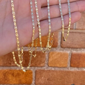 May include: Five gold chains of different lengths, with a textured, diamond-like pattern. The chains are displayed on a hand against a brick wall background.