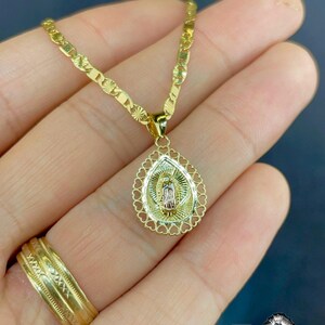 Puede incluir: Collar dorado con colgante religioso. El colgante ovalado tiene un borde en forma de coraz&oacute;n. El collar se muestra en una mano, con un anillo dorado. La cadena es de tipo gourmeta plana. El colgante presenta una figura religiosa.