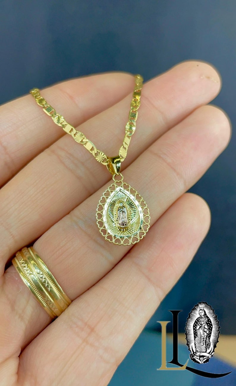Puede incluir: Collar dorado con colgante religioso. El colgante ovalado tiene un borde en forma de coraz&oacute;n. El collar se muestra en una mano, con un anillo dorado. La cadena es de tipo gourmeta plana. El colgante presenta una figura religiosa.