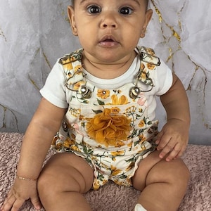 May include: A baby girl wearing a white floral romper with a yellow flower accent. She is sitting on a pink fuzzy rug and wearing white shoes with a pink bow.