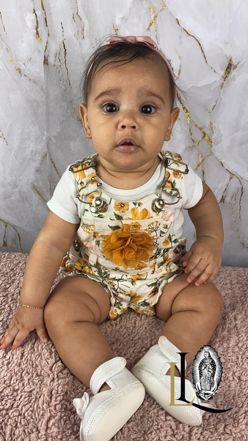 May include: A baby girl wearing a white floral romper with a yellow flower accent. She is sitting on a pink fuzzy rug and wearing white shoes with a pink bow.