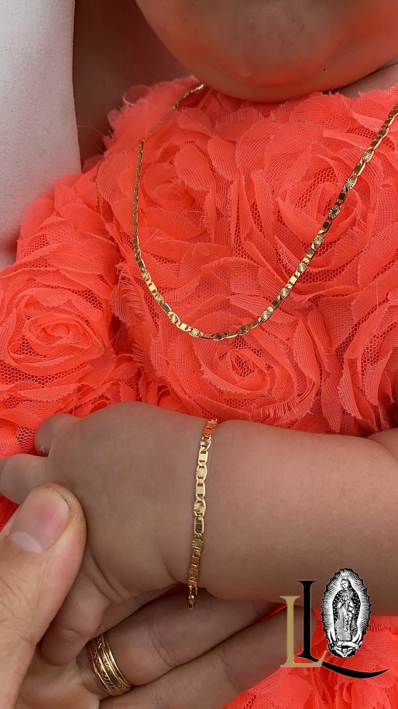 Puede incluir: Un conjunto de collar y pulsera de color dorado. El collar es una cadena delicada con un patr&oacute;n repetitivo, y la pulsera tiene un dise&ntilde;o similar. Las joyas se muestran en una persona que lleva un vestido color coral.