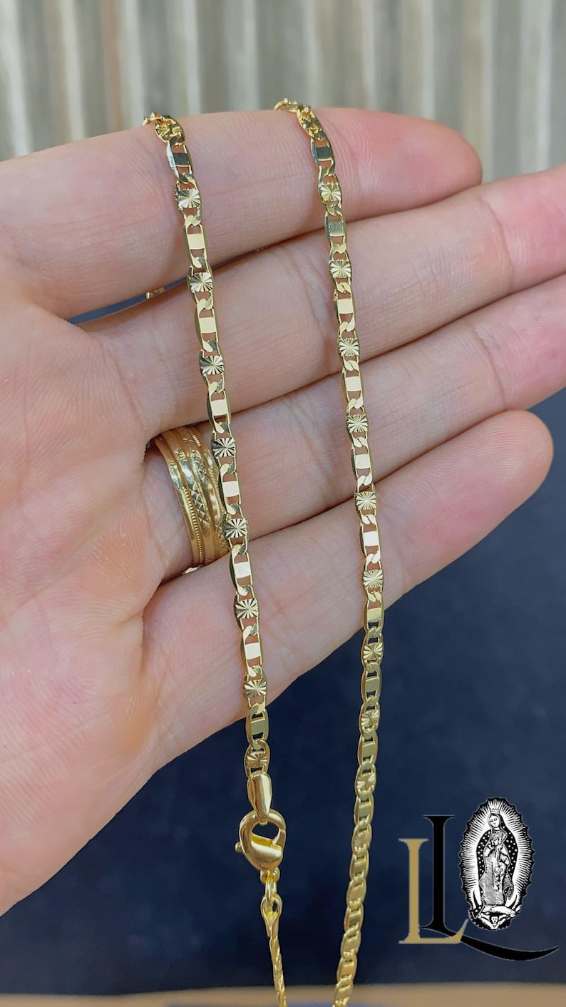 May include: A gold-colored chain necklace with a rectangular link design and starburst accents. The necklace is held in a hand, showcasing its intricate details and the secure clasp. The jewelry is a delicate accessory.