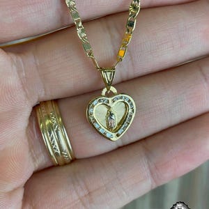 May include: Gold necklace with a heart-shaped pendant, featuring a religious figure and small, clear stones. The chain and pendant have detailed patterns. A gold ring is also visible. The necklace and ring are made of gold.