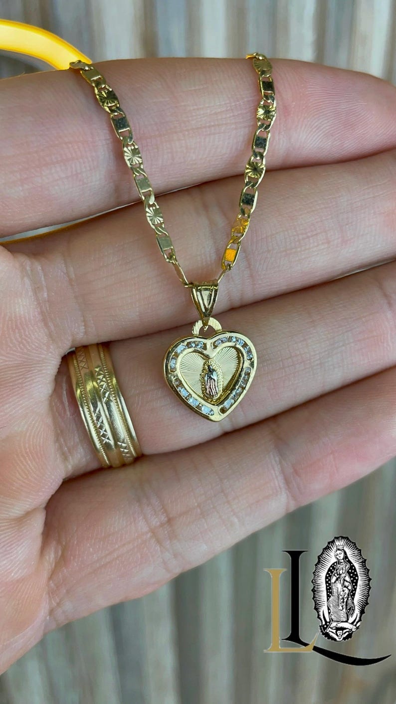 May include: Gold necklace with a heart-shaped pendant, featuring a religious figure and small, clear stones. The chain and pendant have detailed patterns. A gold ring is also visible. The necklace and ring are made of gold.