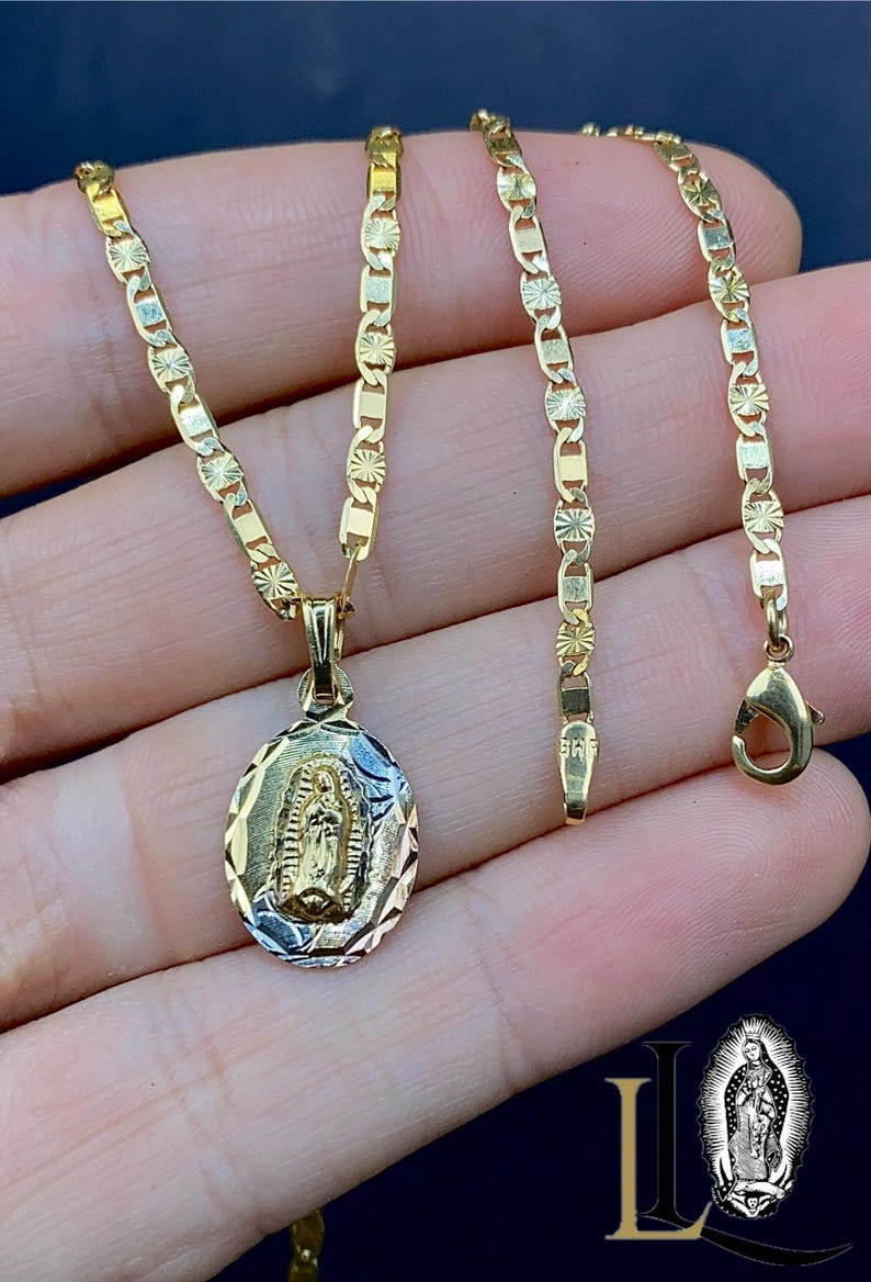 May include: A gold chain necklace with a pendant featuring a depiction of the Virgin Mary. The chain is a Figaro style with alternating links. The pendant is oval shaped and has a detailed design.
