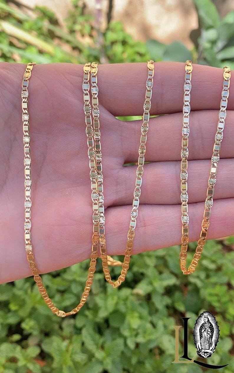 May include: A gold chain necklace with a delicate, patterned design. The chain is made of multiple strands and is shown draped over a hand.