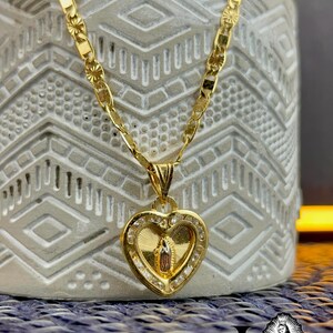 May include: A gold-coloured necklace with a heart-shaped pendant. The pendant features a religious figure and is encrusted with small, clear stones. The chain has a decorative pattern. The necklace is displayed on a textured, grey surface.
