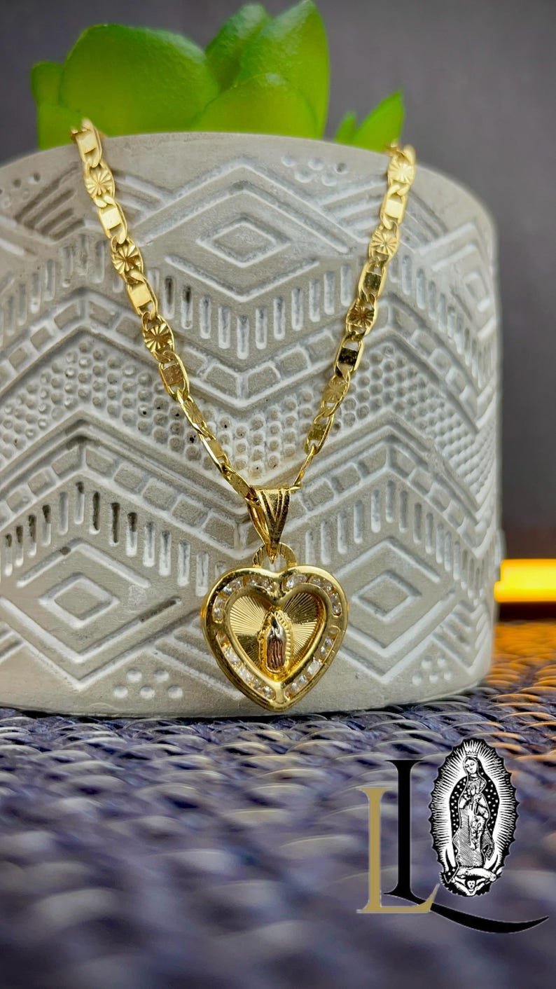 May include: A gold-coloured necklace with a heart-shaped pendant. The pendant features a religious figure and is encrusted with small, clear stones. The chain has a decorative pattern. The necklace is displayed on a textured, grey surface.
