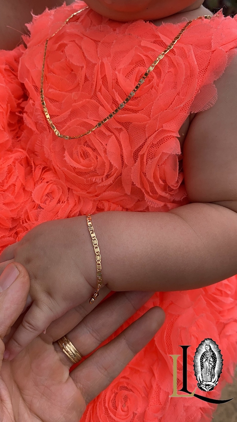 Puede incluir: Un conjunto de collar y pulsera de color dorado. Las joyas est&aacute;n en el brazo de un ni&ntilde;o, con un vestido de color coral brillante en el fondo. Una mano sostiene la mano del ni&ntilde;o. Las joyas parecen estar hechas de peque&ntilde;os eslabones rectangulares.