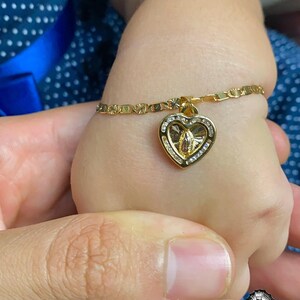 May include: Gold bracelet with a heart-shaped pendant. The pendant features a religious figure and is set with small, clear stones. The bracelet is on a child's wrist, with a parent's hand visible. The background is a blue fabric with white polka dots.