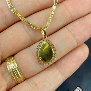 Puede incluir: Un collar dorado con un colgante en forma de l&aacute;grima. El colgante tiene un borde de filigrana y una superficie lisa y reflectante. La cadena es de estilo eslab&oacute;n delicado. Tambi&eacute;n se ve un anillo de oro.