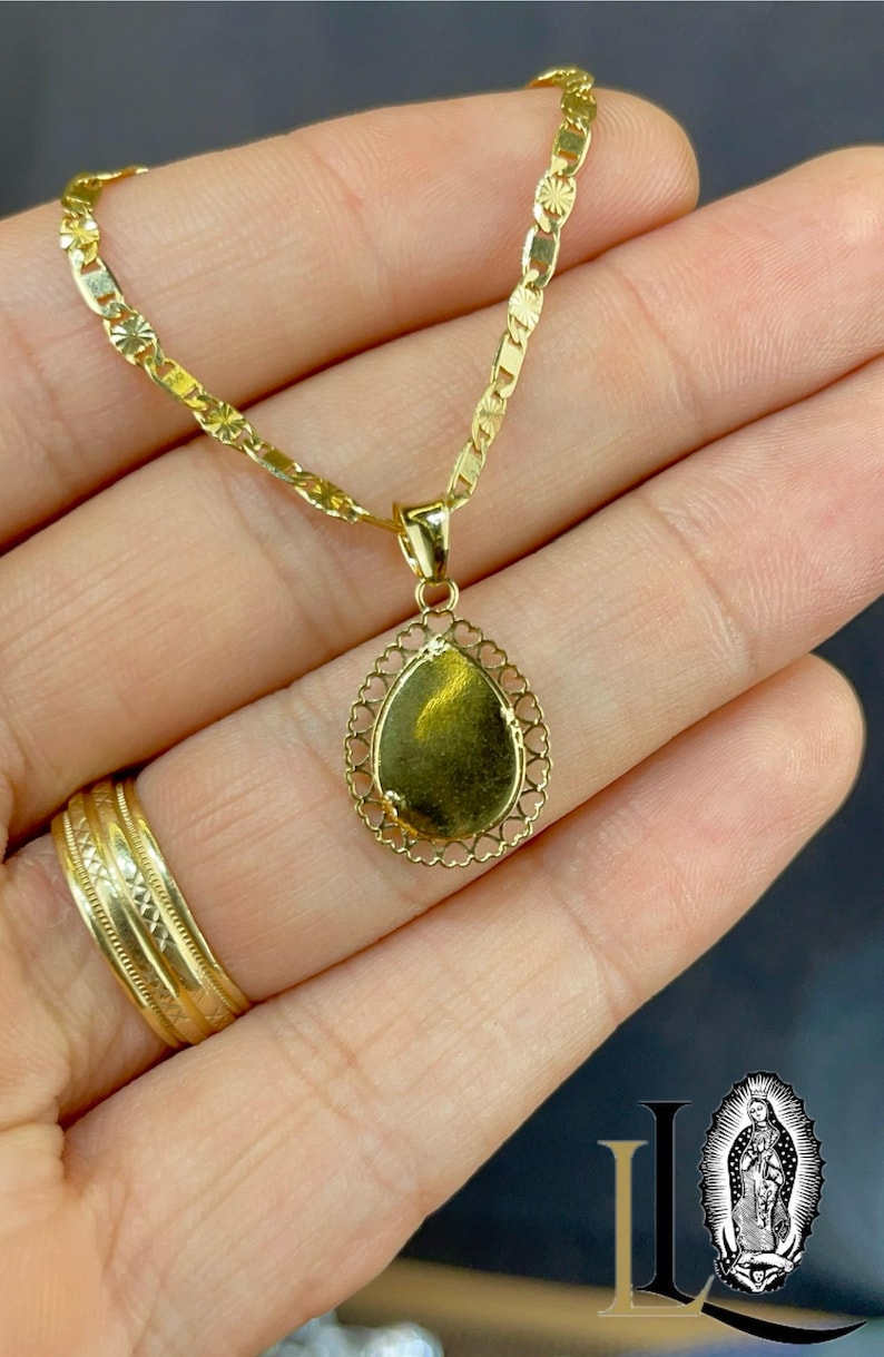 Puede incluir: Un collar dorado con un colgante en forma de l&aacute;grima. El colgante tiene un borde de filigrana y una superficie lisa y reflectante. La cadena es de estilo eslab&oacute;n delicado. Tambi&eacute;n se ve un anillo de oro.