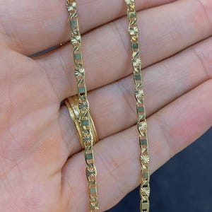 May include: A gold-colored chain necklace with a repeating pattern of rectangular and star-shaped links. The necklace is displayed against a blurred background, with a gold ring visible. The chain is approximately 18 inches long.