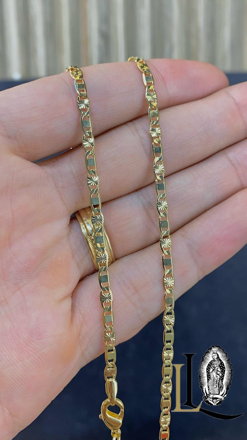 May include: A gold-colored chain necklace with a repeating pattern of rectangular and star-shaped links. The necklace is displayed against a blurred background, with a gold ring visible. The chain is approximately 18 inches long.