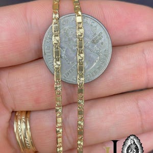 May include: Close-up of two gold-colored chain necklaces with a rectangular and starburst design. The chains are held next to a quarter coin for size comparison. A gold ring is visible on a finger.