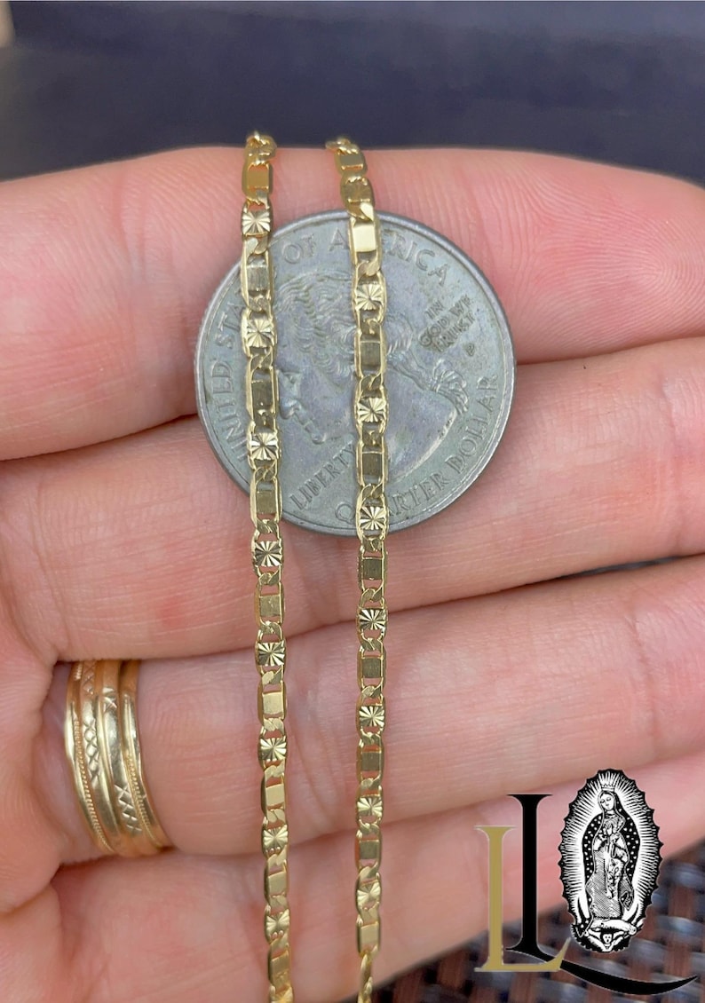 May include: Close-up of two gold-colored chain necklaces with a rectangular and starburst design. The chains are held next to a quarter coin for size comparison. A gold ring is visible on a finger.