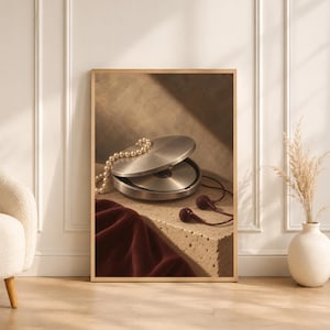 May include: A framed wall art print featuring a silver portable CD player with a string of white pearls draped over the lid. Dark red wired earbuds rest on a textured beige surface next to a folded piece of deep burgundy fabric in soft lighting.