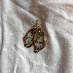 May include: A pair of gold-colored earrings with a unique design. Each earring features a clear, irregular-shaped pendant with a clock face design, including Roman numerals and hands. The earrings hang from silver-colored ear wires.