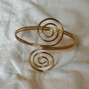 Op de afbeelding: Goudkleurige armmanchet met twee spiraalvormige ontwerpen. De manchet heeft een dubbelbandontwerp dat om de arm gewikkeld is. De spiralen zijn gecentreerd op de manchet, waardoor een decoratief accessoire ontstaat. De manchet ligt op een witte ondergrond.