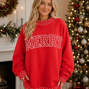 May include: A red long-sleeve sweatshirt featuring the word MERRY in white block letters with a polka dot pattern. The garment includes a mock neck collar, sleeve cuffs, and a bottom hem, all accented with a white polka dot pattern on a red background.