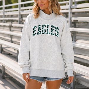 May include: A light gray heathered long-sleeve sweatshirt with a mock neck collar. The front features the word EAGLES in a dark green collegiate font with white polka dots. The sweatshirt has dropped shoulders and ribbed cuffs at the wrists and hem.