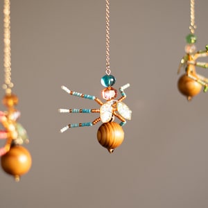 Puede incluir: Tres adornos decorativos de insectos colgando de cadenas doradas. Cada insecto presenta un cuerpo de madera, patas con cuentas y alas iridiscentes. Las cuentas son en tonos de azul, rosa y verde. El fondo es gris neutro.