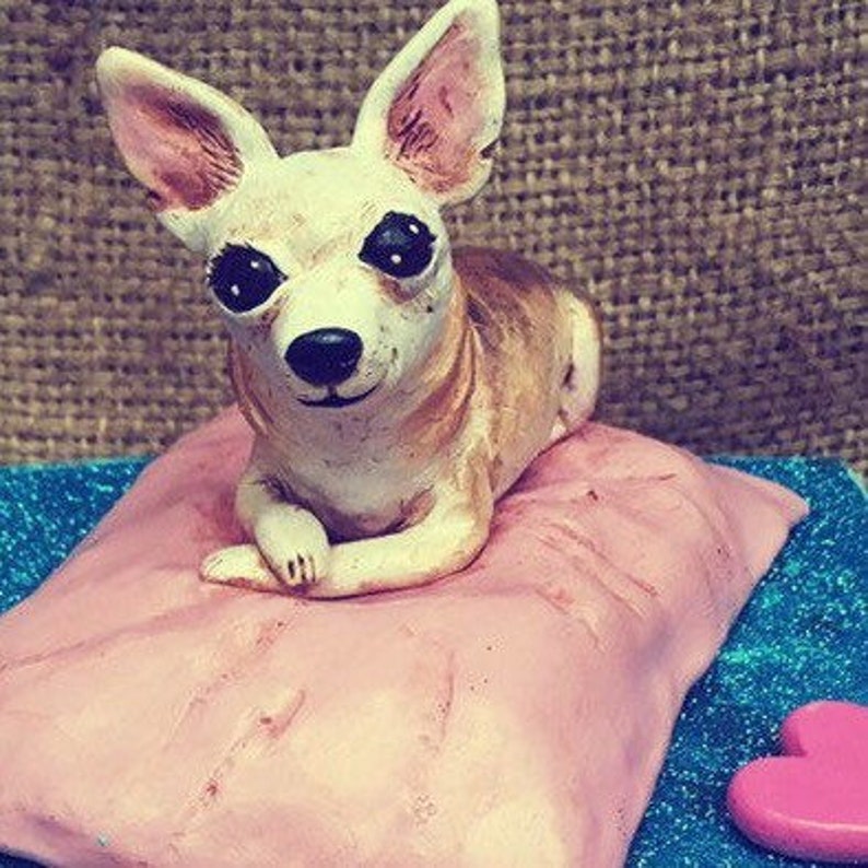May include: A small, white ceramic chihuahua figurine with black eyes, sitting on a pink pillow. The figurine is on a blue and silver glitter background.