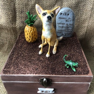 May include: A brown wooden box with a glitter lid. The box has a small metal latch and a nameplate that reads "Pi&ntilde;a". The lid features a small ceramic figurine of a chihuahua dog, a pineapple, and a small green lizard.