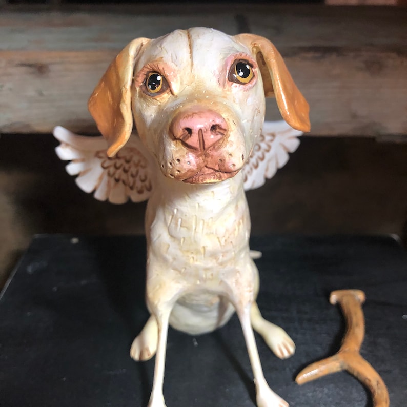 May include: A white ceramic dog figurine with brown ears and wings. The dog has a sweet expression and is sitting with its legs outstretched.