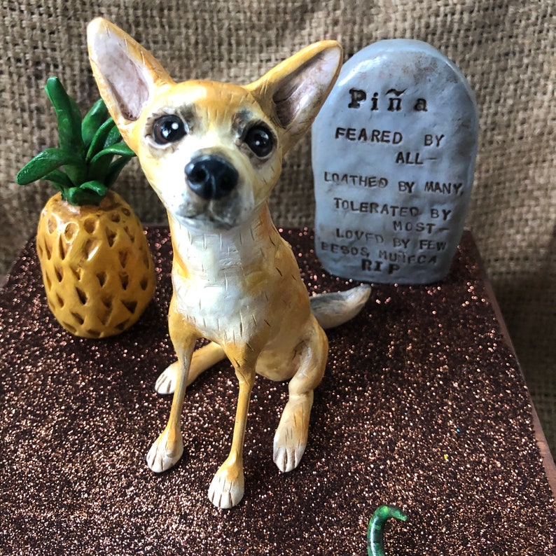 May include: A ceramic figurine of a tan chihuahua dog with a tombstone that reads "Pi&ntilde;a Feared By All - Loathed By Many Tolerated By Most Loved By Few Besos Munifica RIP".