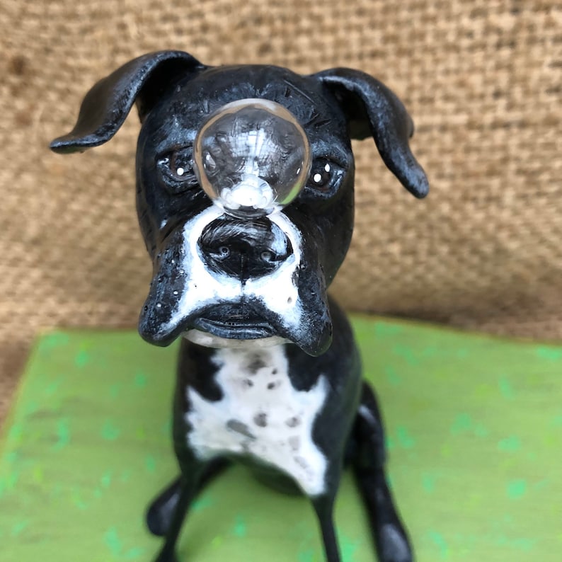 May include: A black and white boxer dog figurine with a clear glass bubble on its nose. The figurine is sitting on a green surface.
