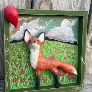 Könnte beinhalten: Gerahmtes Kunstwerk mit einem Fuchs mit orangefarbenem Fell und weißen Akzenten, der einen roten Ballon hält. Der Hintergrund zeigt ein Feld roter Blumen und einen bewölkten Himmel. Der grüne Rahmen hat eine Juteaufhängung. Ca. 20 cm.