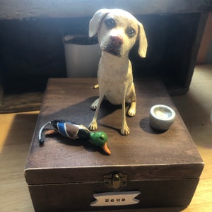 May include: A white Labrador Retriever figurine with a brown wooden box, a green and blue duck decoy, and a silver metal bowl. The box has a brass latch and a name tag that reads "Zeus".