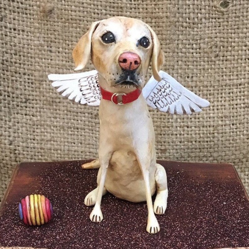 May include: A ceramic sculpture of a yellow Labrador Retriever with white wings, wearing a red collar. The dog is sitting on a brown surface with a rainbow-colored ball in front of it.