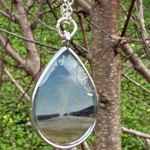 May include: Silver-toned teardrop pendant necklace. The pendant features a landscape scene reflected within the glass. The chain is a delicate silver link. The necklace hangs against a backdrop of tree branches and green foliage.