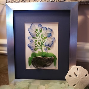 May include: A framed artwork featuring a blue flower made of sea glass, with green leaves and a gray shell base. The artwork is surrounded by a silver frame and a blue mat.