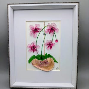 May include: Framed artwork featuring a handcrafted orchid arrangement. The pink flowers with dark centers are set against a white background. The pot is a light brown color. The frame is white with a gray wood-grain texture.