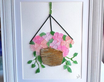 May include: A framed artwork featuring a hanging flower pot made of sea glass. The pot is filled with pink and white flowers and green leaves. The artwork is on a white background.