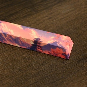 May include: A rectangular keyboard keycap featuring a scenic landscape design. The keycap displays a mountain range with a pagoda, rendered in shades of purple, pink, and orange, evoking a sunset or sunrise. The keycap is set against a dark wooden surface.