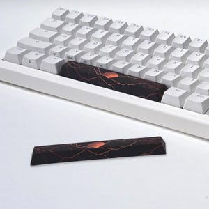 May include: A white mechanical keyboard featuring a custom black spacebar with a graphic of a red moon rising over dark mountain silhouettes with orange outlines. An identical loose spacebar is positioned in the foreground for comparison.