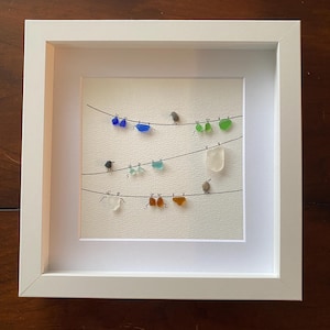 May include: A framed artwork with three horizontal lines adorned with colourful glass pieces and small bird figures. The glass pieces are in shades of blue, green, orange, and white. The artwork is set in a white frame.