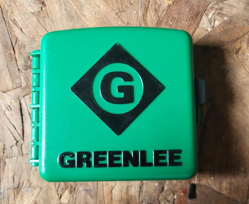 Op de afbeelding: Een felgroene, vierkante gereedschapskist met de merknaam "GREENLEE" in het wit. Het heeft een zwarte ruit met een witte "G". De kist heeft een scharnierzijde en een sluiting.