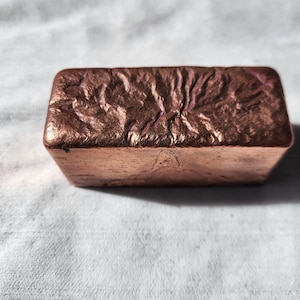 May include: A copper-coloured rectangular object with a textured, wrinkled surface. The object has a metallic sheen and appears solid. The edges are clean and defined, contrasting with the textured top. It is likely a decorative item or a small sculpture.