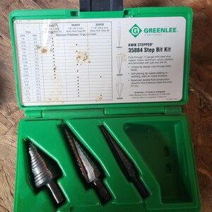 May include: A green plastic case containing three black step drill bits. The case has a label with size and measurement information in inches and millimeters. The label also includes the text "KWIK STEPPER 35884 Step Bit Kit".