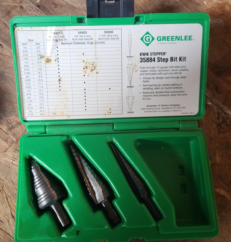 Op de afbeelding: Een groene plastic koffer met drie zwarte trapboren. De koffer heeft een label met maat- en meetinformatie in inches en millimeters. Het label bevat ook de tekst "KWIK STEPPER 35884 Step Bit Kit".