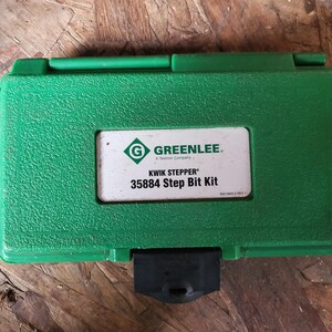 Op de afbeelding: Een groene plastic gereedschapskist met een getextureerd oppervlak. De kist heeft een wit label met de tekst "GREENLEE" en "KWIK STEPPER 35884 Step Bit Kit". De kist is gesloten en heeft een zwarte sluiting.