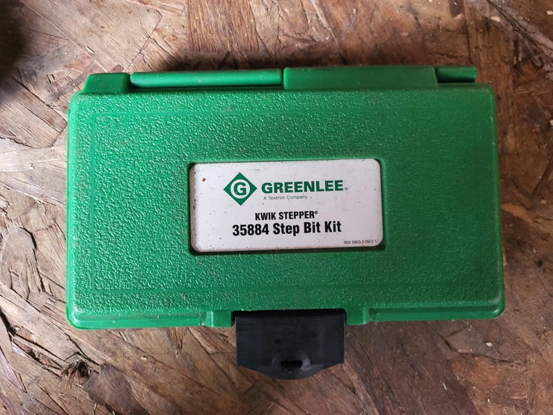 Op de afbeelding: Een groene plastic gereedschapskist met een getextureerd oppervlak. De kist heeft een wit label met de tekst "GREENLEE" en "KWIK STEPPER 35884 Step Bit Kit". De kist is gesloten en heeft een zwarte sluiting.