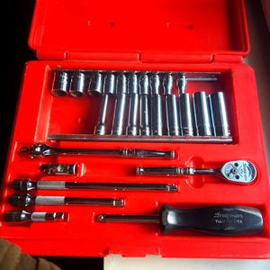 Peut inclure: Une boîte à outils rouge contenant un assortiment de douilles chromées, de cliquets et de rallonges. Les outils sont rangés dans le boîtier ouvert. Un tournevis Snap-on TM-124 USA noir et argent est également visible.