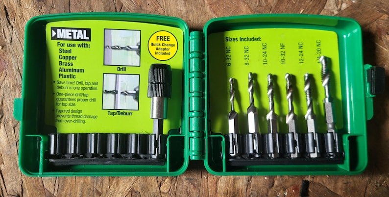 Op de afbeelding: Open groene plastic koffer met een set boren en een snelwisseladapter. De koffer toont verschillende boormaten voor metaal en kunststof. De tekst op de koffer geeft aan dat de boren geschikt zijn voor staal, koper, messing, aluminium en kunststof.